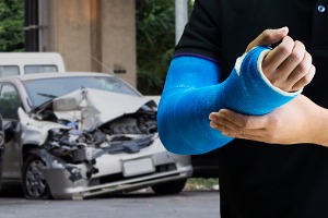 A man with a broken arm in front of a crashed car, needing an Auto Accident Attorney in Peoria IL